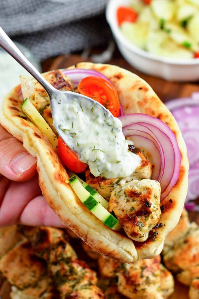 pieces of grilled chicken, fresh tomatoes, onion, and cucumbers sit inside a piece of pita bread held by a hand. More pieces of chicken and vegetables can be seen in the background e