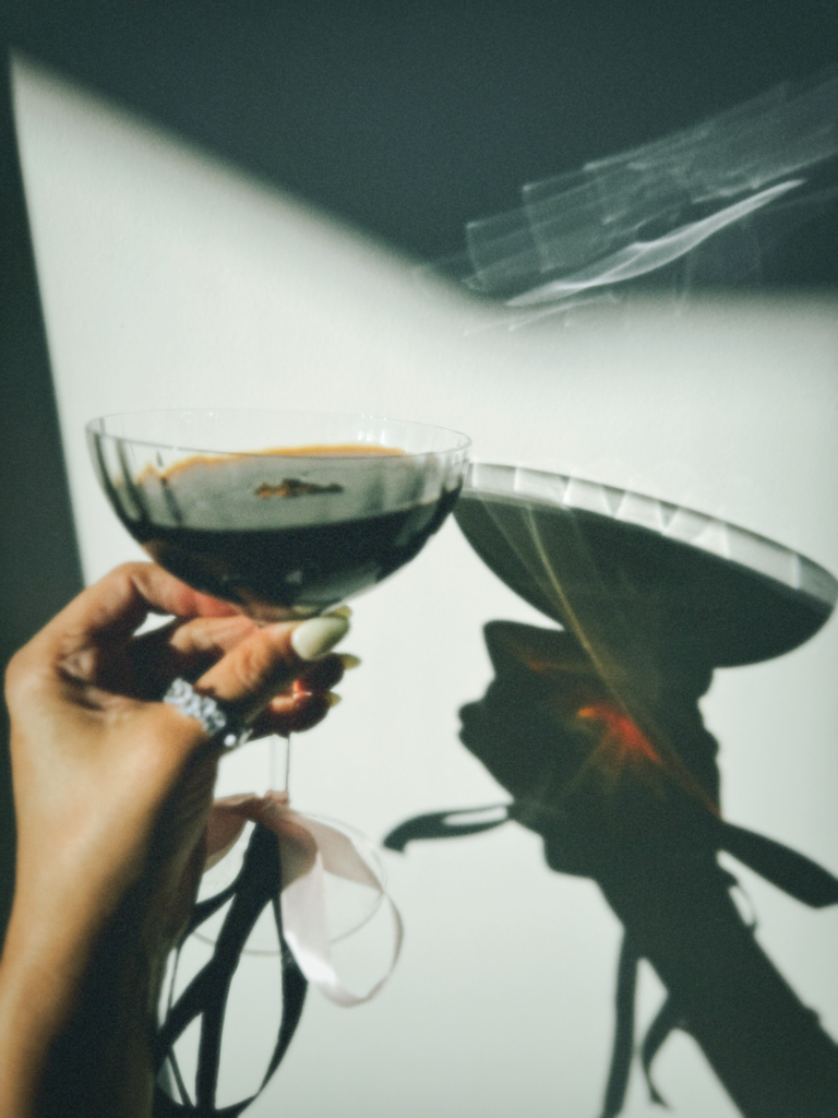A hand holds a dark cocktail in front of a white wall where the reflection of the cocktail can been. The cocktail is close to the wall appearing to toast the reflection. 